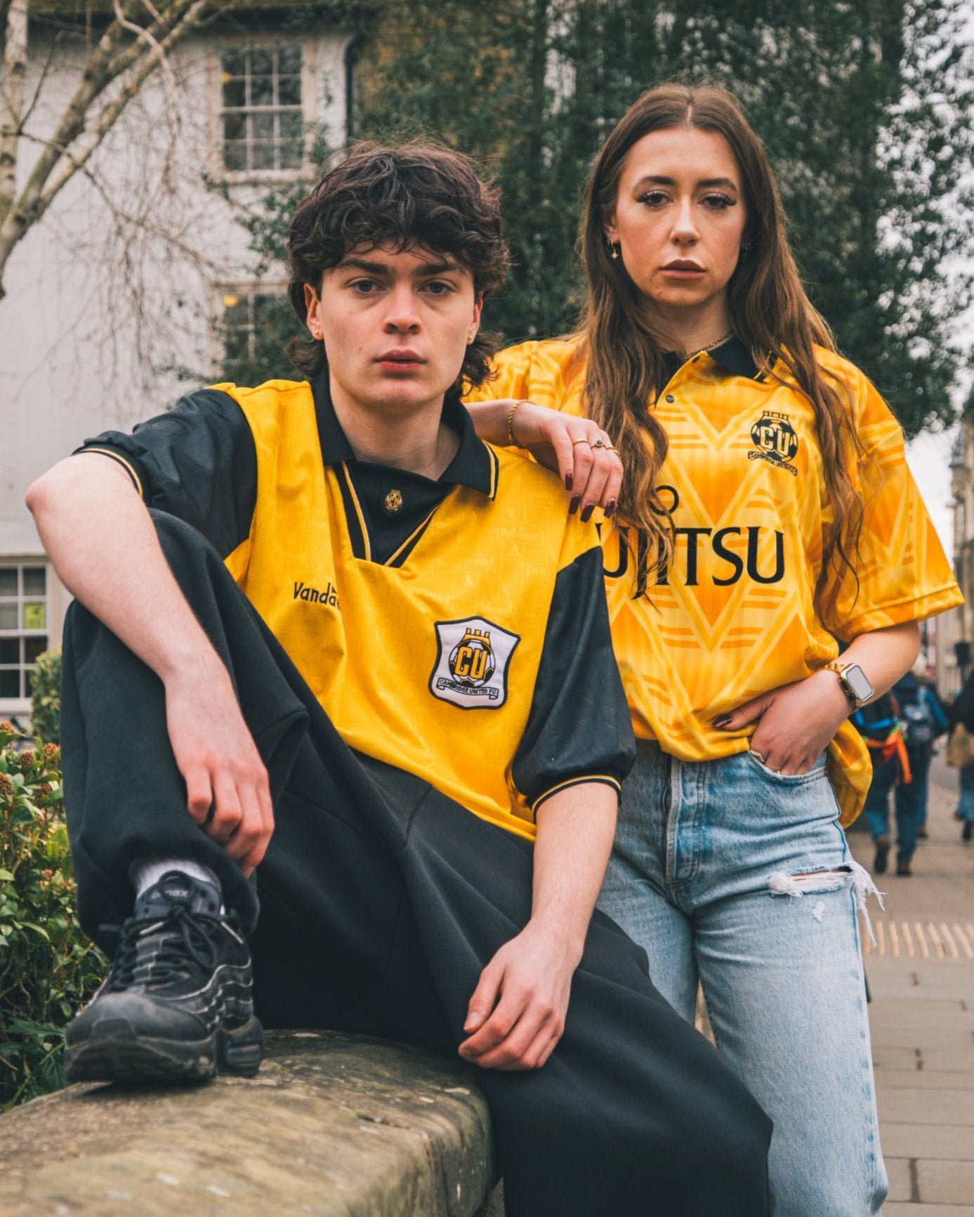 Two people wearing yellow sports jerseys sitting outdoors with a building and trees in the background.