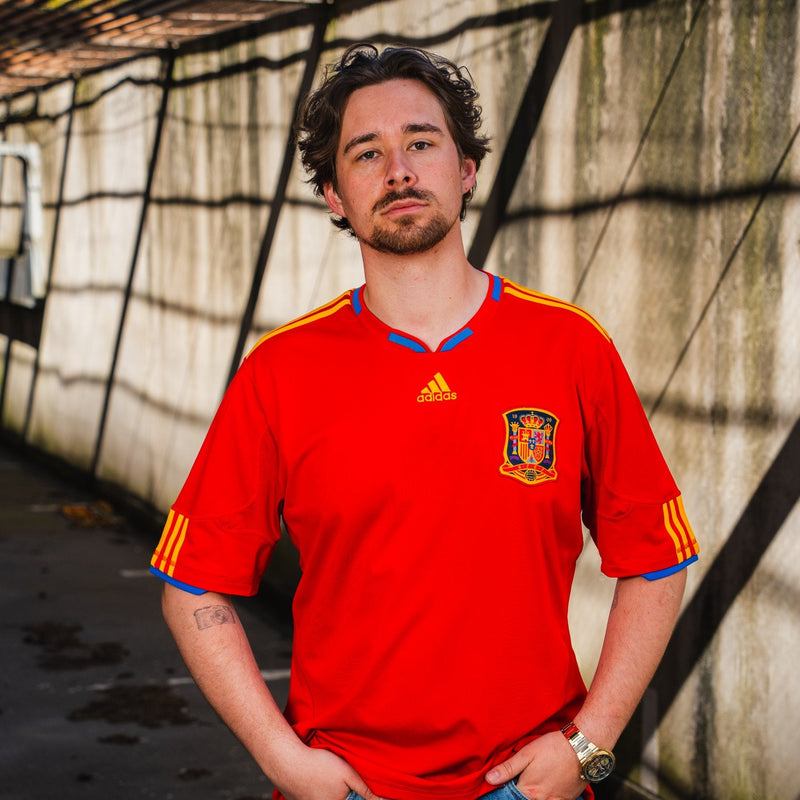 Person wearing a red Spain soccer jersey with Adidas logo against a textured wall.