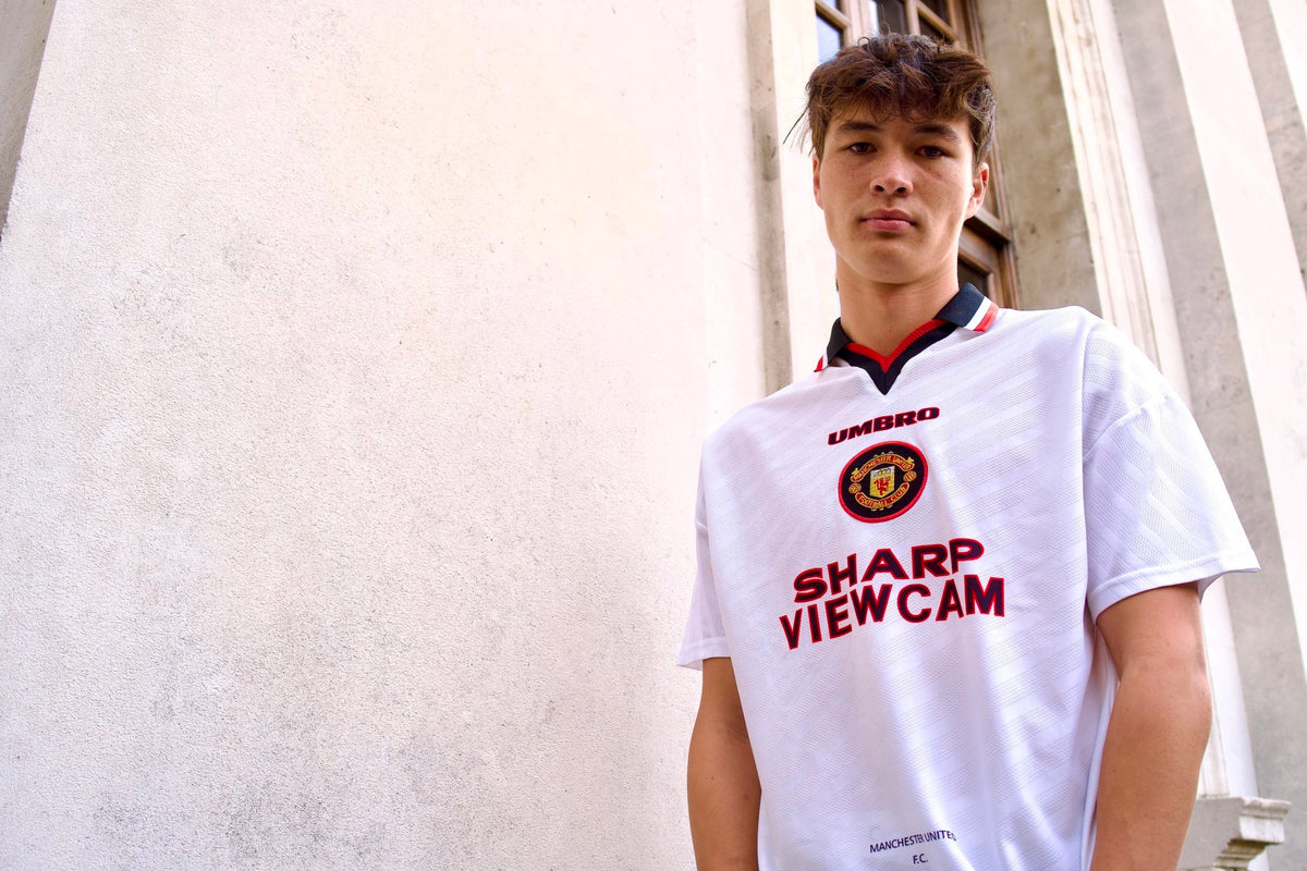A young man wearing a white Manchester United Umbro football jersey with red and navy trim, standing against a light-colored stone wall.