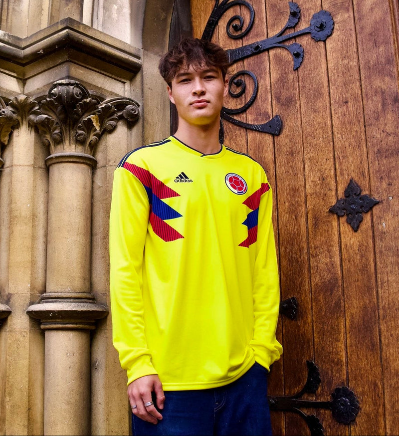 A young man wearing a bright yellow long-sleeve soccer jersey with Colombian national team graphics and an Adidas logo, standing in front of a historic stone column and wooden door.
