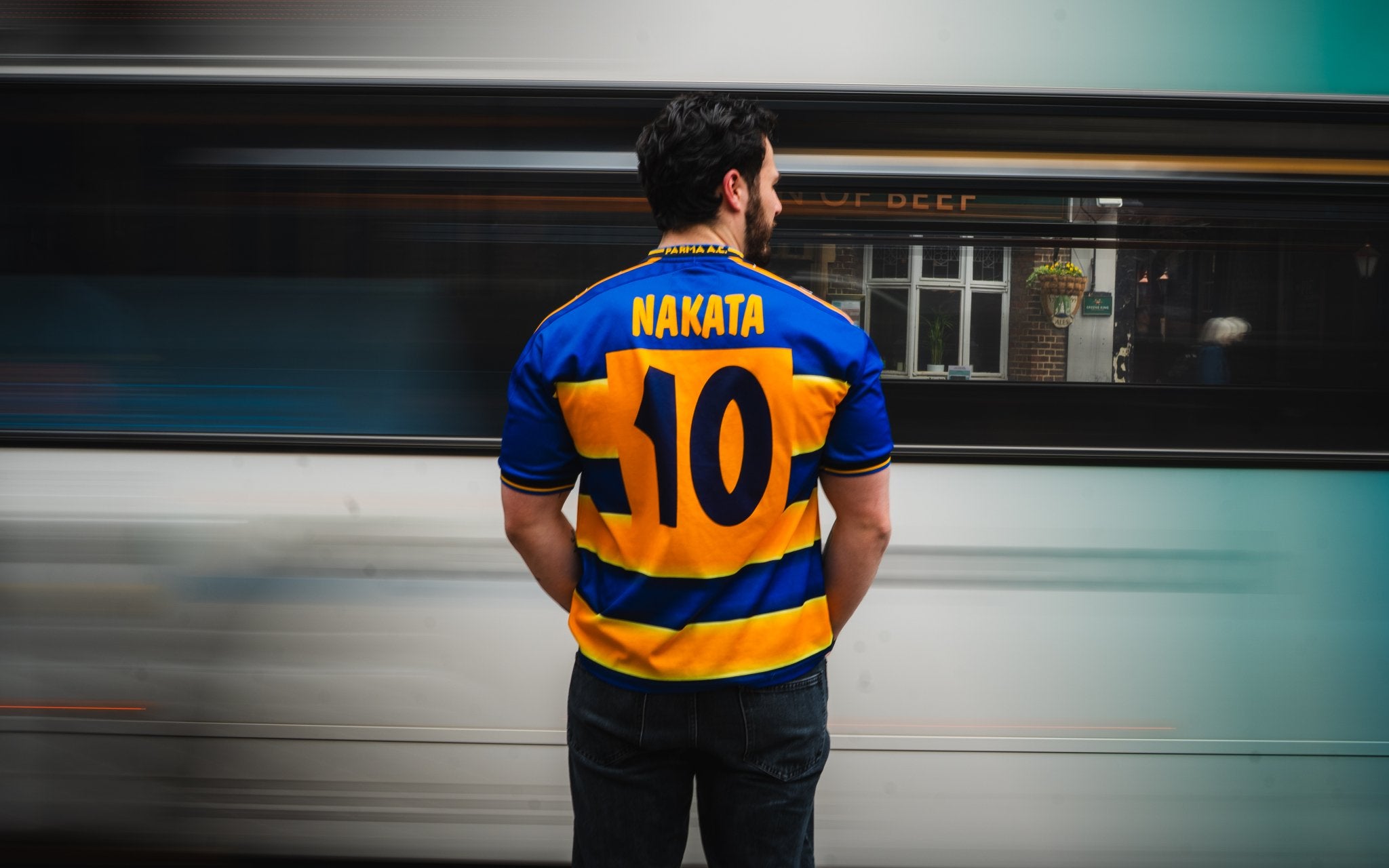 Person wearing a blue and yellow sports jersey with 'Nakata 10' on the back, standing in front of a moving train.