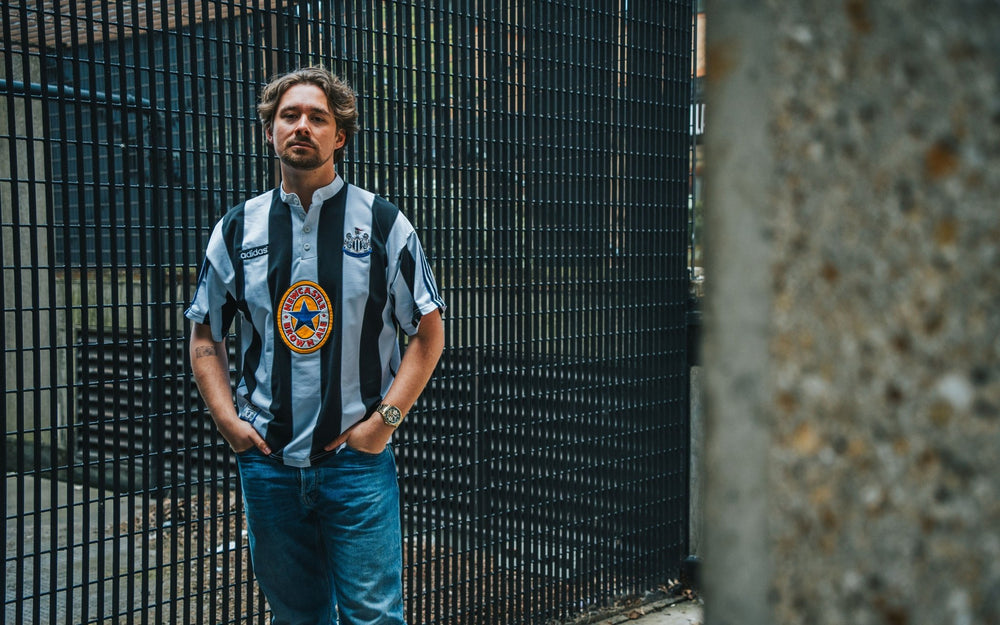 Person wearing a black and white striped shirt with a logo, standing against a metal fence.