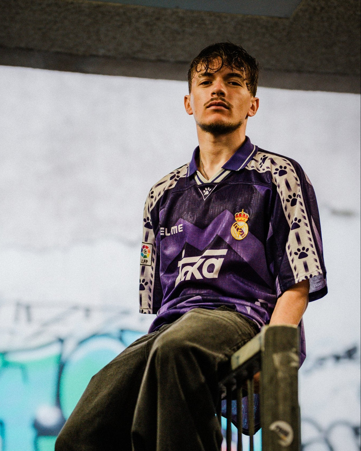 A man wearing a purple and white Real Madrid soccer jersey with 'FLYE' and 'FCB' logos, paired with dark gray baggy pants, sitting on a metal railing in front of a graffiti wall.