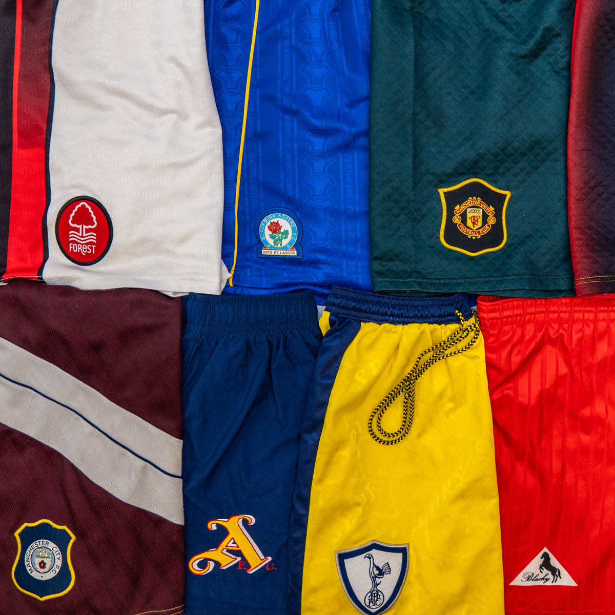 A flat-lay grid of multiple athletic shorts in different colors, including white, blue, green, maroon, navy, yellow, and red, each showing a team crest or logo patch.